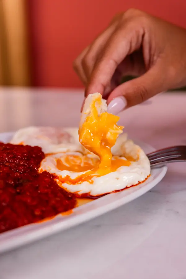Mano mojando pan en la yema de un huevo frito con tomate